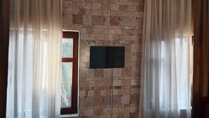 A room with a brick wall, a flat-screen TV mounted on it, and sheer curtains on the windows. - Mylos Otel (Bozcaada)