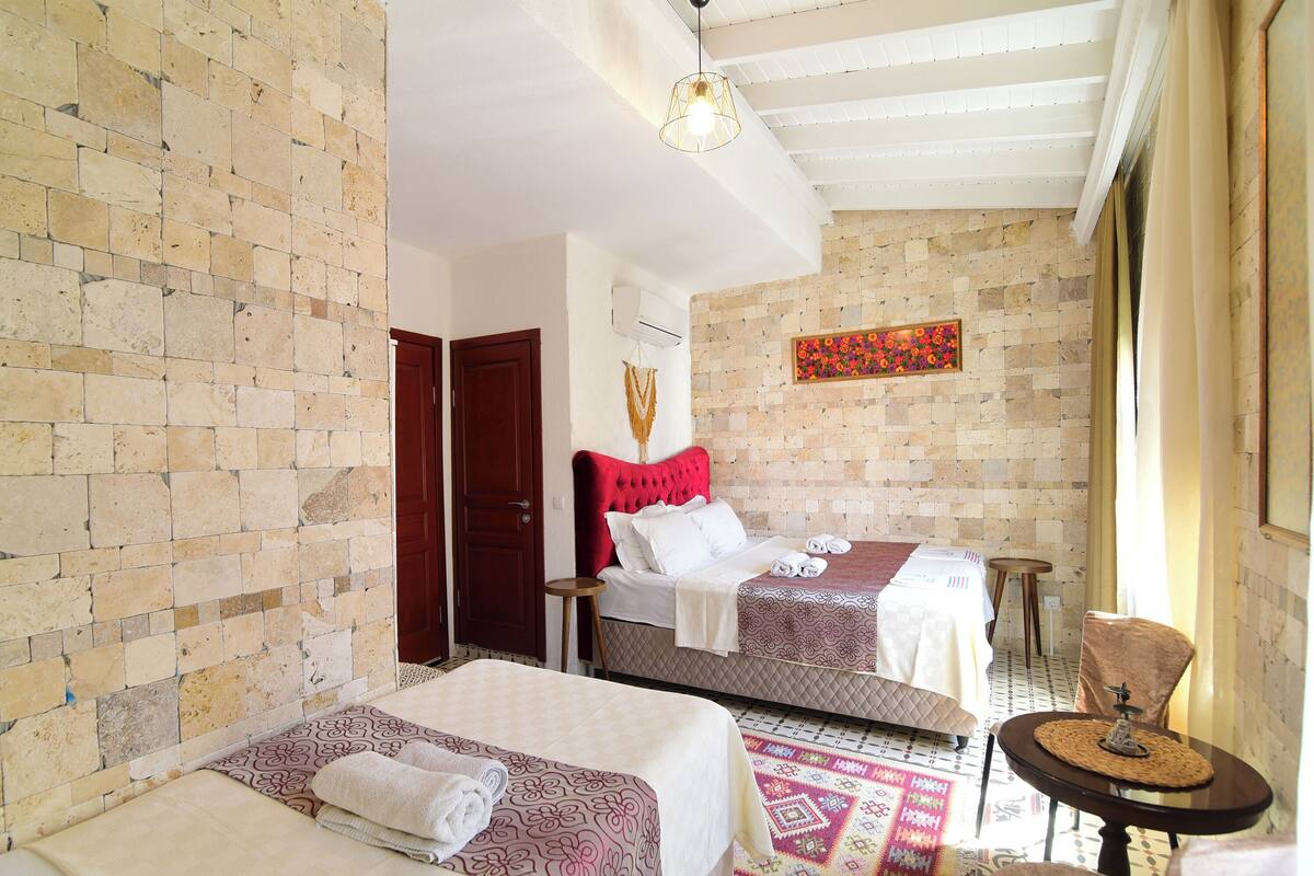 Habitación de hotel con dos camas, cabecera roja, alfombra estampada y una pequeña mesa redonda con un objeto decorativo.