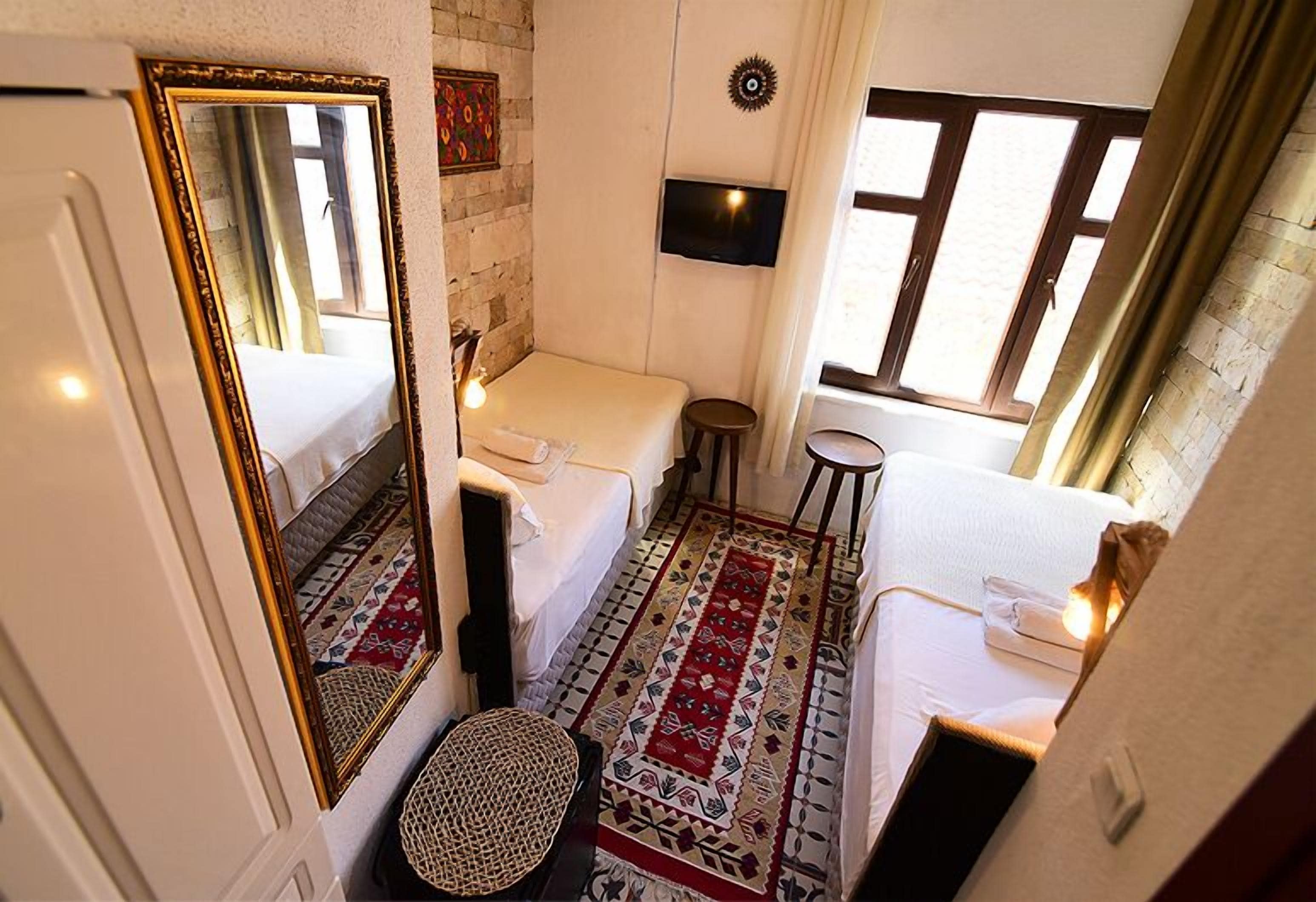 A compact hotel room with a single bed, a small stool, a mirror, a television, and a window with curtains.