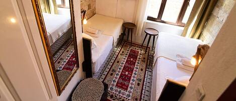 A compact hotel room with a single bed, a small stool, a mirror, a television, and a window with curtains.