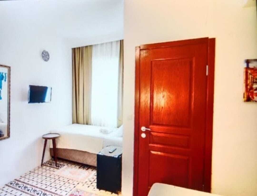 Hotellihuone, jossa on sänky, pieni pöytä, tuoli, ikkuna verhoilla ja puinen ovi.