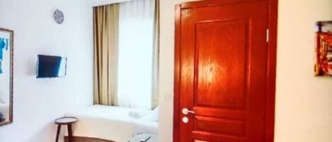 A hotel room with a bed, a small table, a chair, a window with curtains, and a wooden door.