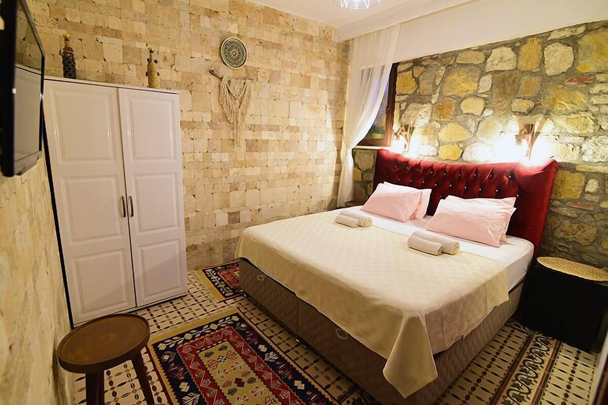 Un dormitorio con pared de piedra, cama con cabecera roja, armario blanco y alfombra estampada.