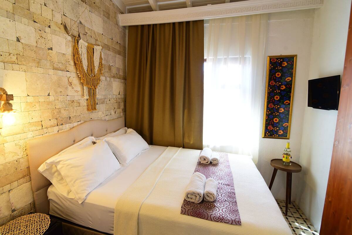 Habitación de hotel con pared de ladrillo, cama con sábanas blancas, cabecera de madera, una mesita redonda con lámpara y un cuadro floral en la pared.