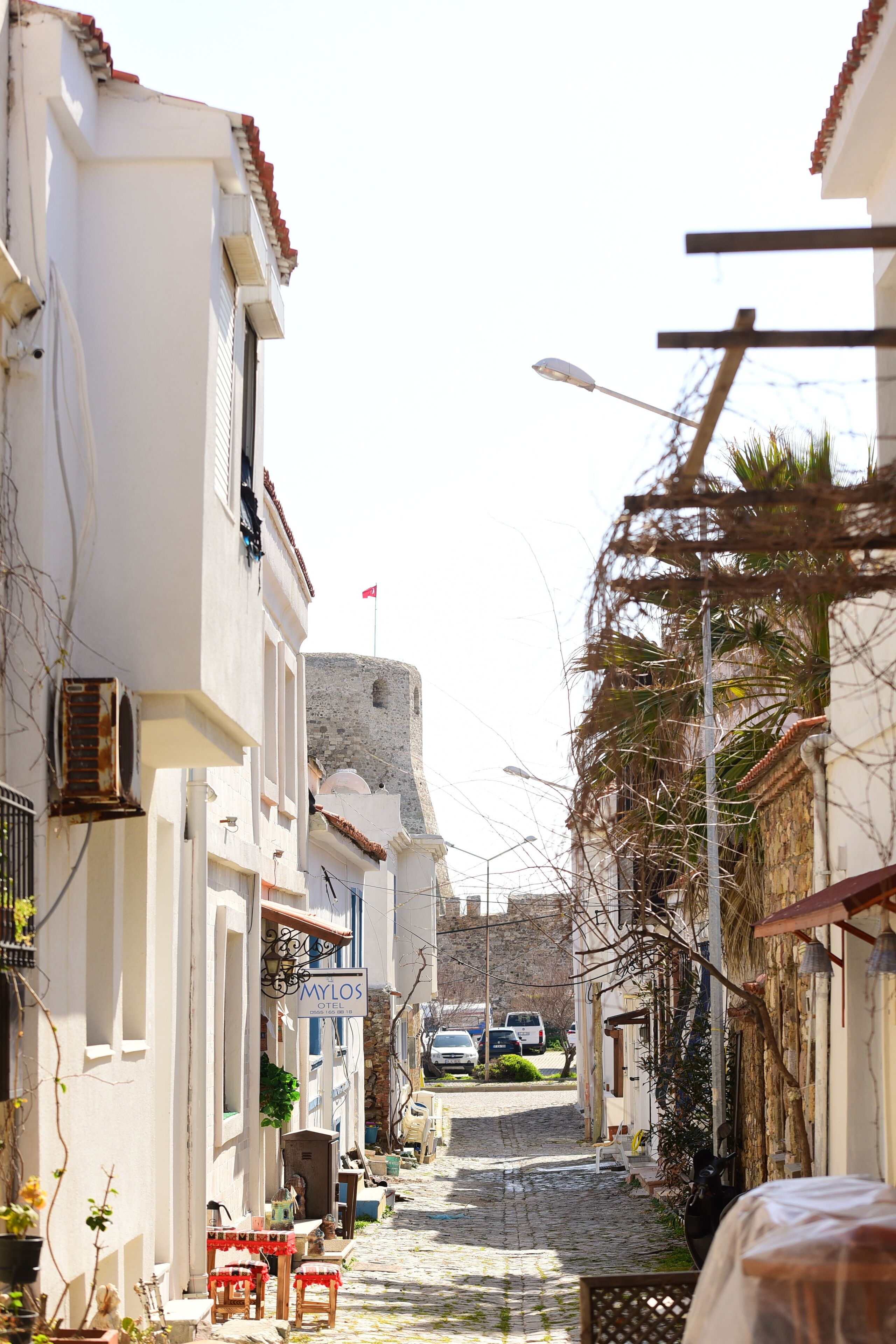 A narrow cobblestone street lined with white buildings, a sign for 'MYLOS HOTEL', and a distant view of a fort.