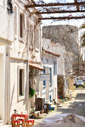 A narrow street with a hotel named "MYLOS HOTEL" on the right. There are outdoor seating areas with tables and chairs. - Mylos Otel (Bozcaada)