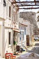 Una calle estrecha con un hotel llamado "MYLOS HOTEL" a la derecha. Se observan zonas de estar al aire libre con mesas y sillas.