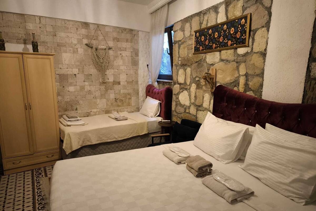 Habitación de hotel con pared de piedra, cama con ropa blanca, armario de madera y un tapiz de macramé en la pared.