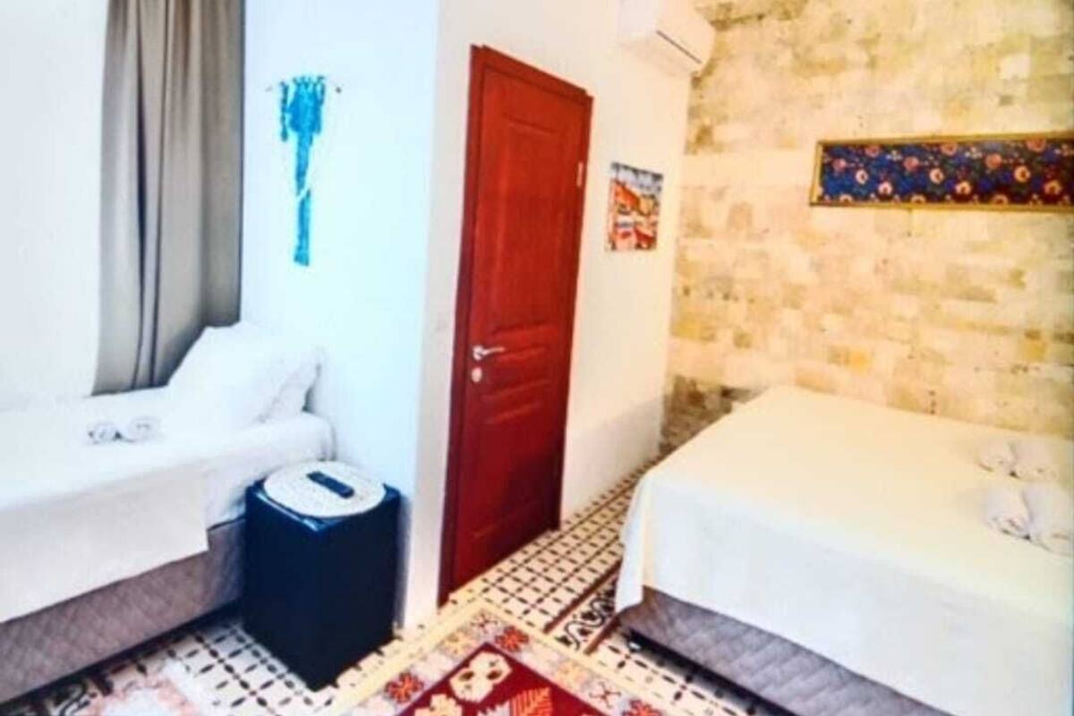Una habitación con cama, una mesita, una silla, un ventanal con cortinas y un aire acondicionado instalado en la pared.