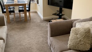 A living room with beige carpet, a beige sofa, a television, and a dining table with chairs. - First floor, recently renovated central Llandudno apartment (Llandudno)