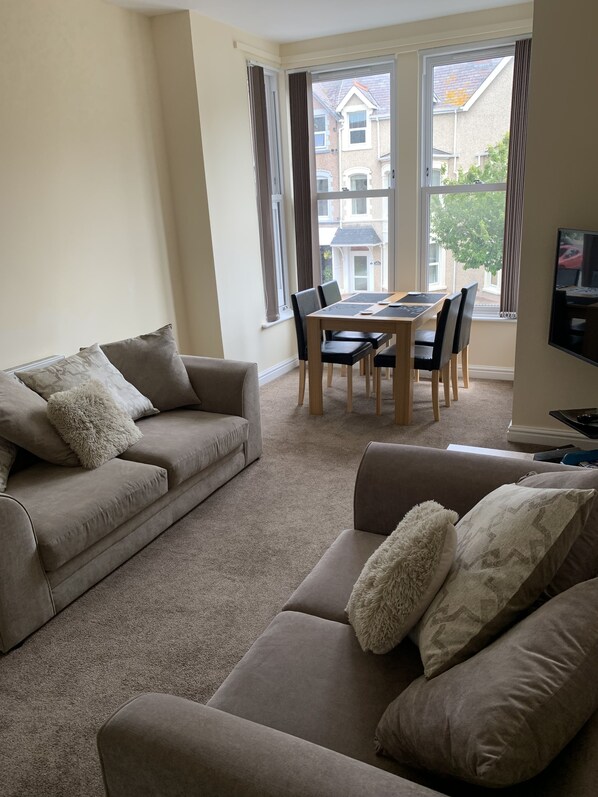 Un salon avec un canapé beige, un canapé plus petit, une table à manger avec des chaises et un téléviseur fixé au mur.