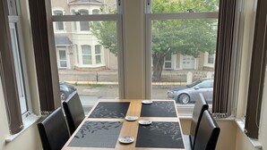 A dining area with a wooden table and four black chairs, large windows with curtains, and a view of neighboring houses. - First floor, recently renovated central Llandudno apartment (Llandudno)