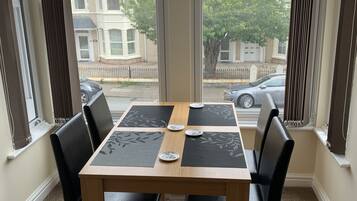 Un coin repas avec une table en bois et quatre chaises noires, de grandes fenêtres avec des rideaux et une vue sur les maisons voisines.