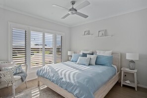 Ein Schlafzimmer mit einem Bett, Deckenventilator, Nachttisch, Sessel und einem Fenster mit Jalousien.