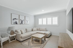 Ein modernes Wohnzimmer mit einem beigefarbenen Ecksofa, einem Holztisch und einem weißen Sessel. An der Wand hängen drei gerahmte Tierbilder und es gibt ein Fenster mit weißen Fensterläden.