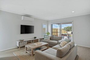 Ein modernes Wohnzimmer mit einem Flachbildfernseher, grauen Sofas, einem Couchtisch und einer Glasschiebetür, die zu einem Außenbereich führt.