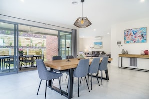 A modern dining area with a wooden table and grey chairs, a hanging light fixture, and a glass door leading to an outdoor patio with plants and a brick wall. - Vue Sur La Mer by Ready Set Stay - View of the sea (Mount Martha)