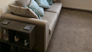 Una sala de estar con un sofá color beige, una mesita con una botella y un objeto decorativo, y una ventana con vistas a casas y vegetación.