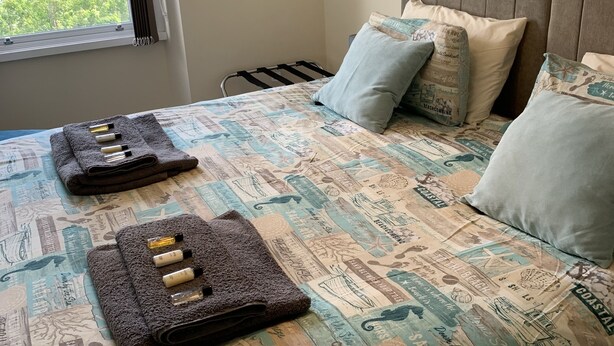 Una cama bien hecha con una colcha de estilo costero, almohadas y una mesita de noche con botellas.