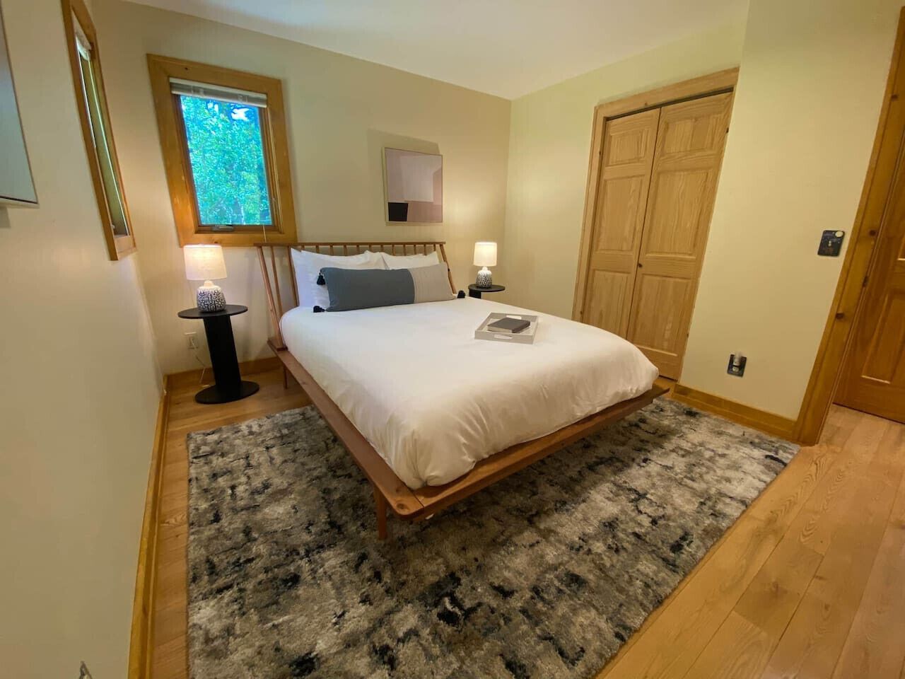 A bedroom with a bed, two bedside lamps, a small table, a window with a view, and a wooden door.