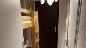 Un pasillo estrecho con una cama litera de madera a la izquierda, una puerta oscura al final y una puerta blanca a la derecha.