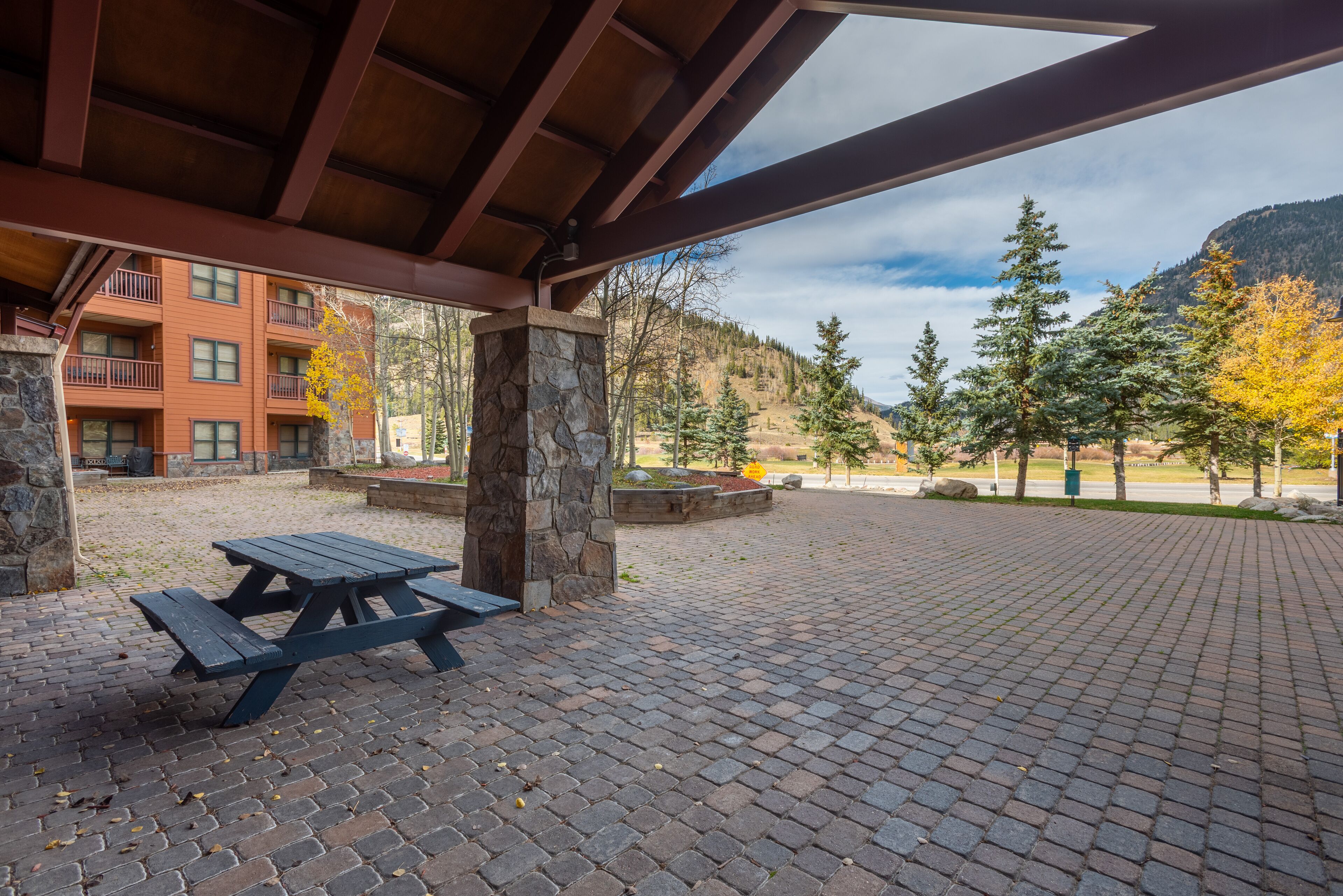 Un espace extérieur abrité, doté d’une table de pique-nique, de piliers en pierre et offrant une vue sur un bâtiment avec des balcons, le tout sur fond de paysage montagneux.