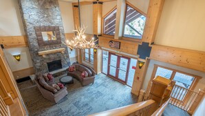 A spacious living area with a stone fireplace, a chandelier, and a large window overlooking a deck. - Right next to Super Bee Lift, wifi, underground parking (Copper Mountain)