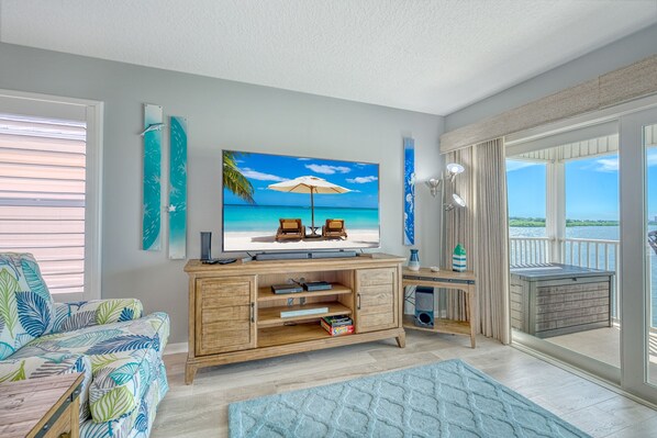 A living room with a large TV, wooden entertainment unit, and a view of the ocean through large windows.