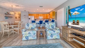 Eine moderne Küche mit Holzausstattung, ein Essbereich mit Holztisch und -stühlen sowie ein Wohnzimmer mit gemusterten Sesseln und einem Fernseher, der eine Strandansicht zeigt.