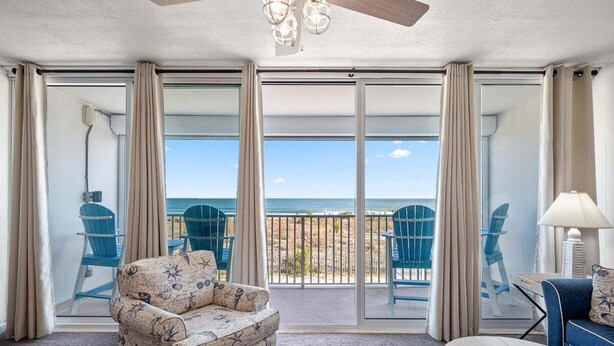 A room with a large sliding glass door leading to a balcony with a view of the beach. There are two blue chairs and a patterned armchair inside the room.