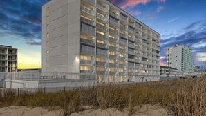 A modern hotel with multiple floors and balconies, situated near a beach with sand and grass.