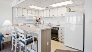 A compact kitchen with white cabinets, a built-in microwave, a refrigerator, and a small dining area with white chairs.