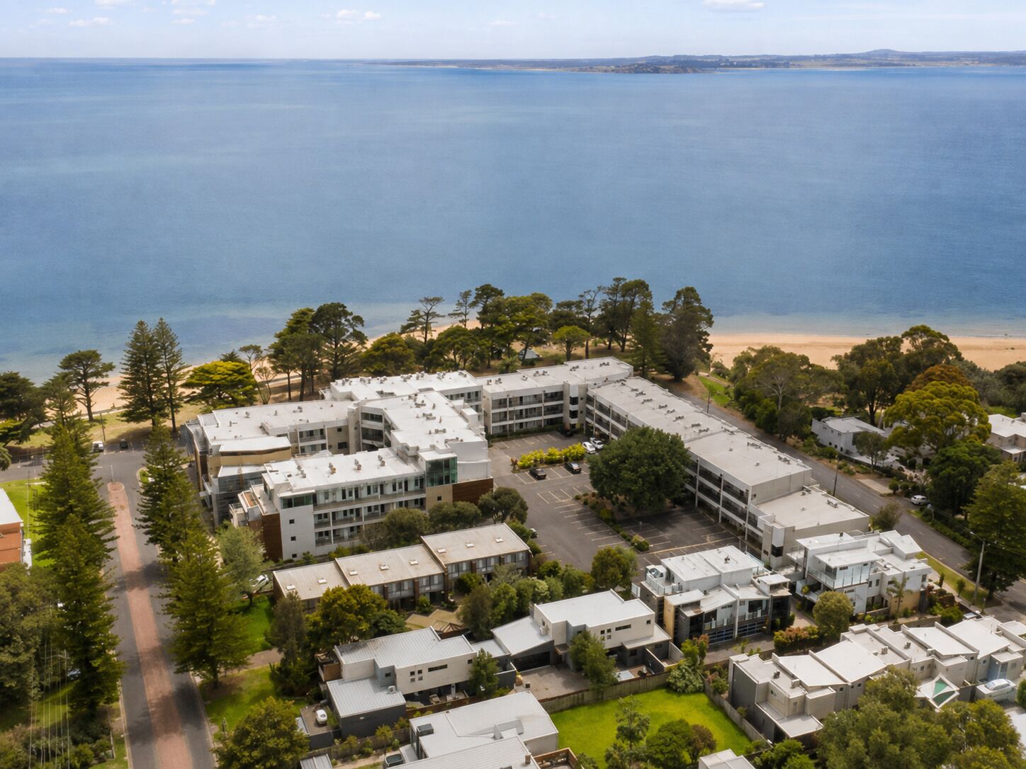 A coastal residential area with modern buildings, a parking lot, and a beach in the background.