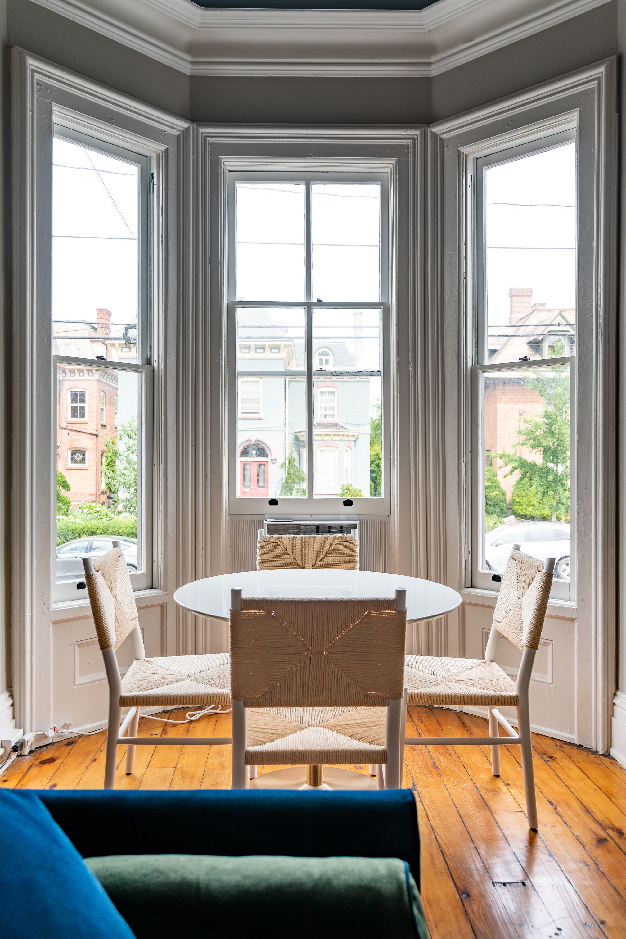 Une baie vitrée composée de trois grands panneaux, une table ronde avec deux chaises, et une vue sur un bâtiment en briques rouges et sur la verdure environnante.