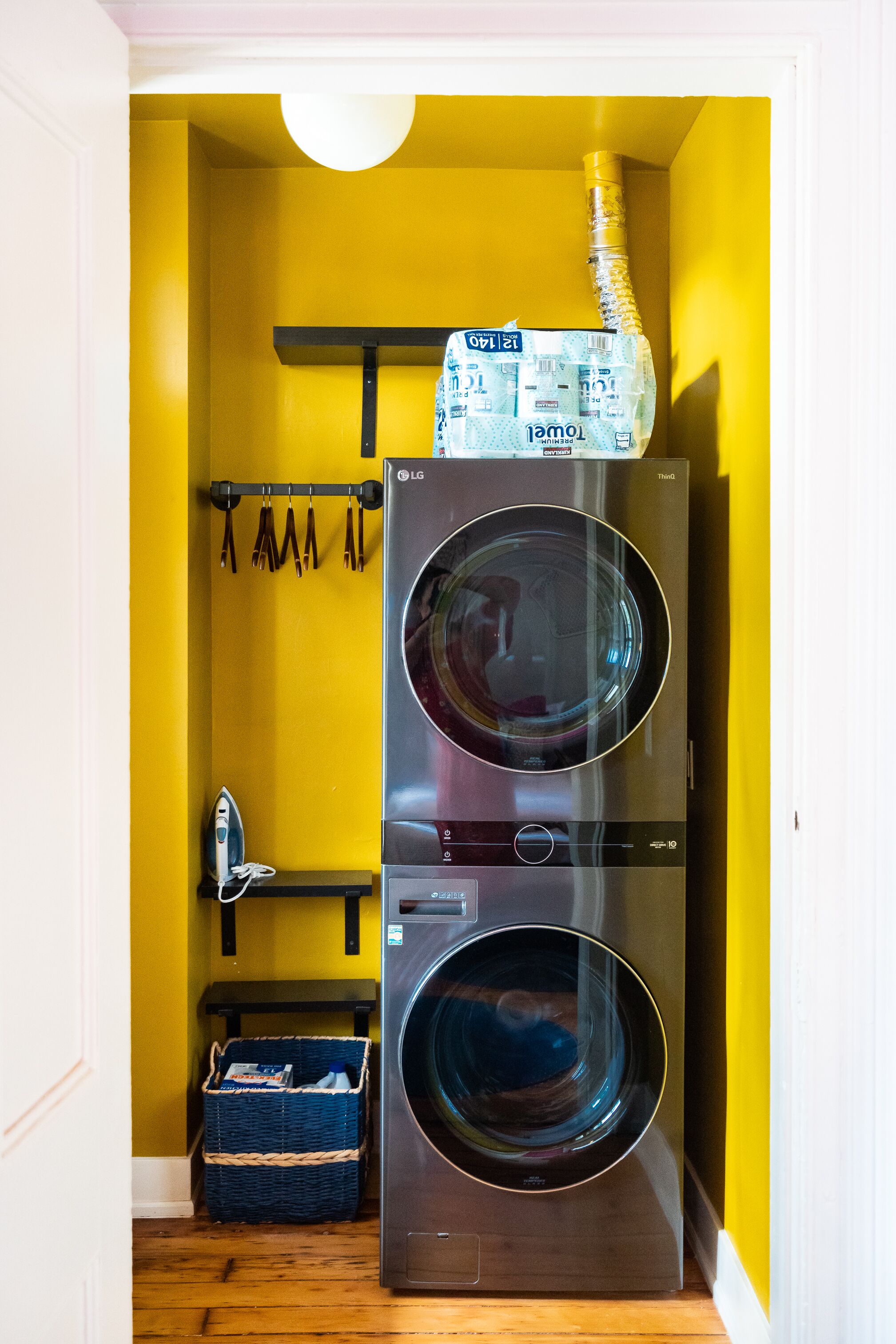 A laundry room with two stacked front-loading washing machines, a drying rack with hanging clothes, and a basket with laundry detergent.