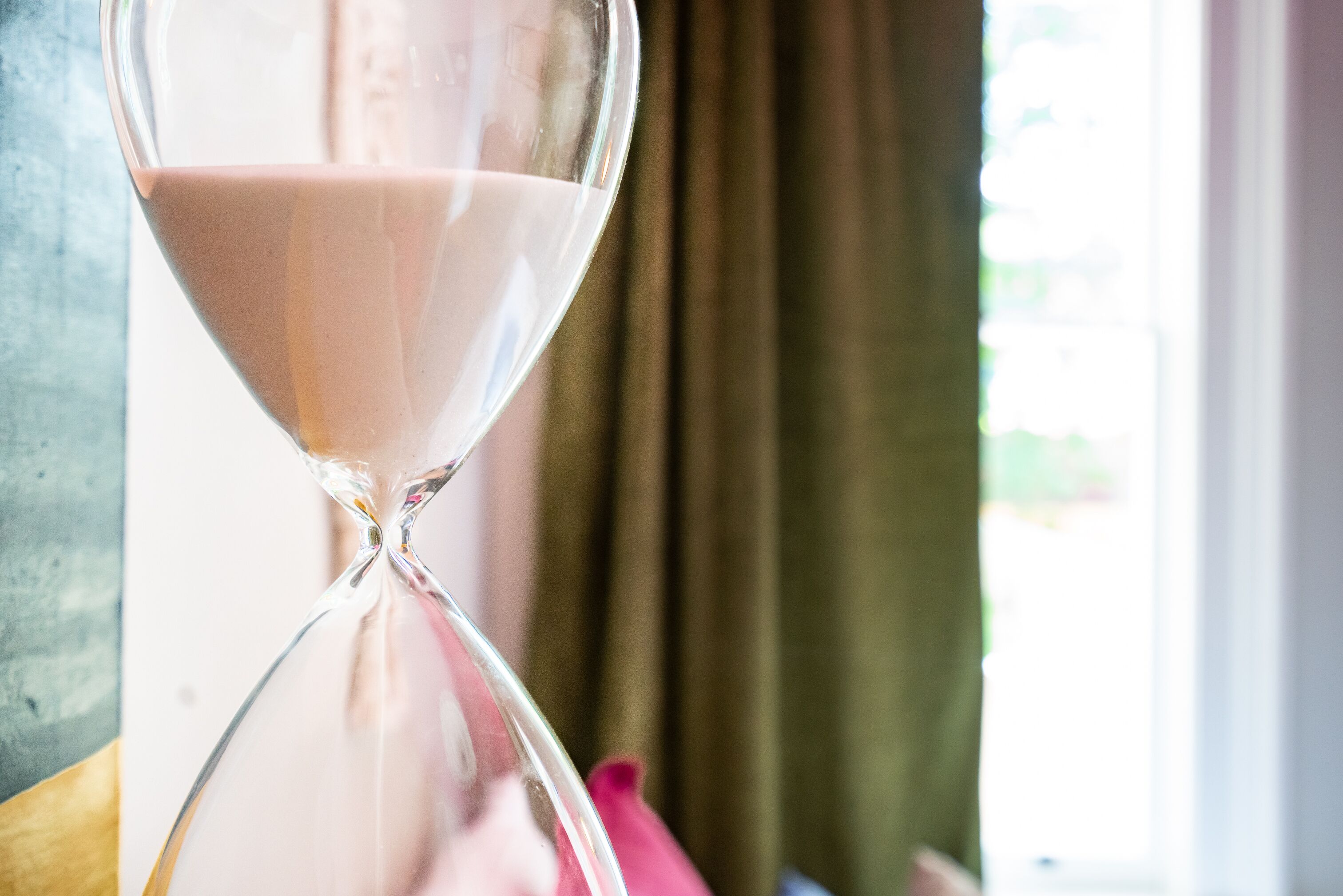 An hourglass with a pink liquid, placed on a surface near a window with curtains.