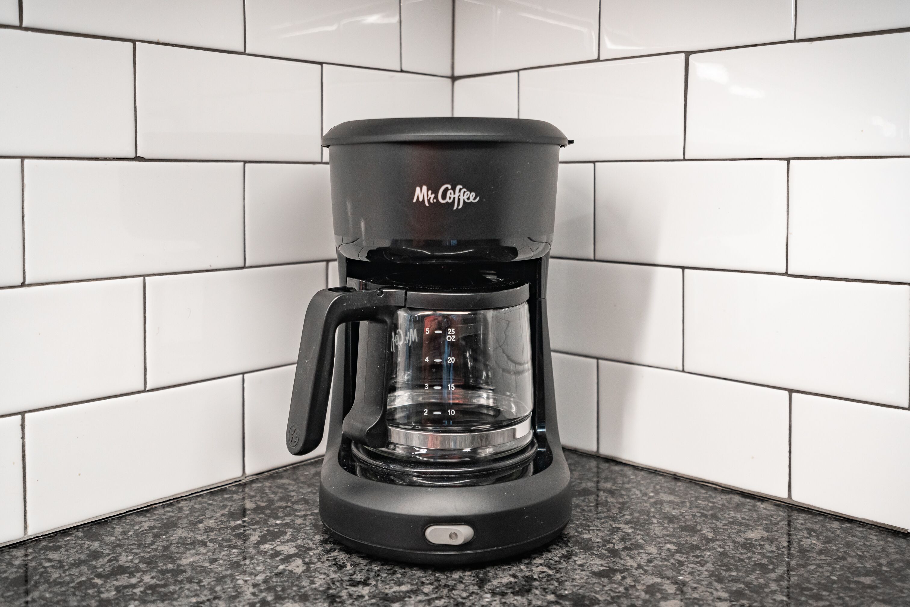 Une machine à café noire de la marque Mr. Coffee, posée sur un comptoir en granit, avec des carreaux de métro blancs en arrière-plan.