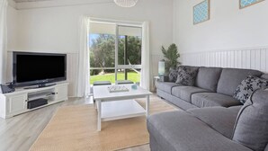 A living room with a flat-screen TV, a grey sofa, a white coffee table, and a large window with a view of greenery. - Seaesta Rye by Ready Set Stay - Pet-friendly, solar heated pool, WIFI, Netflix (Rye)