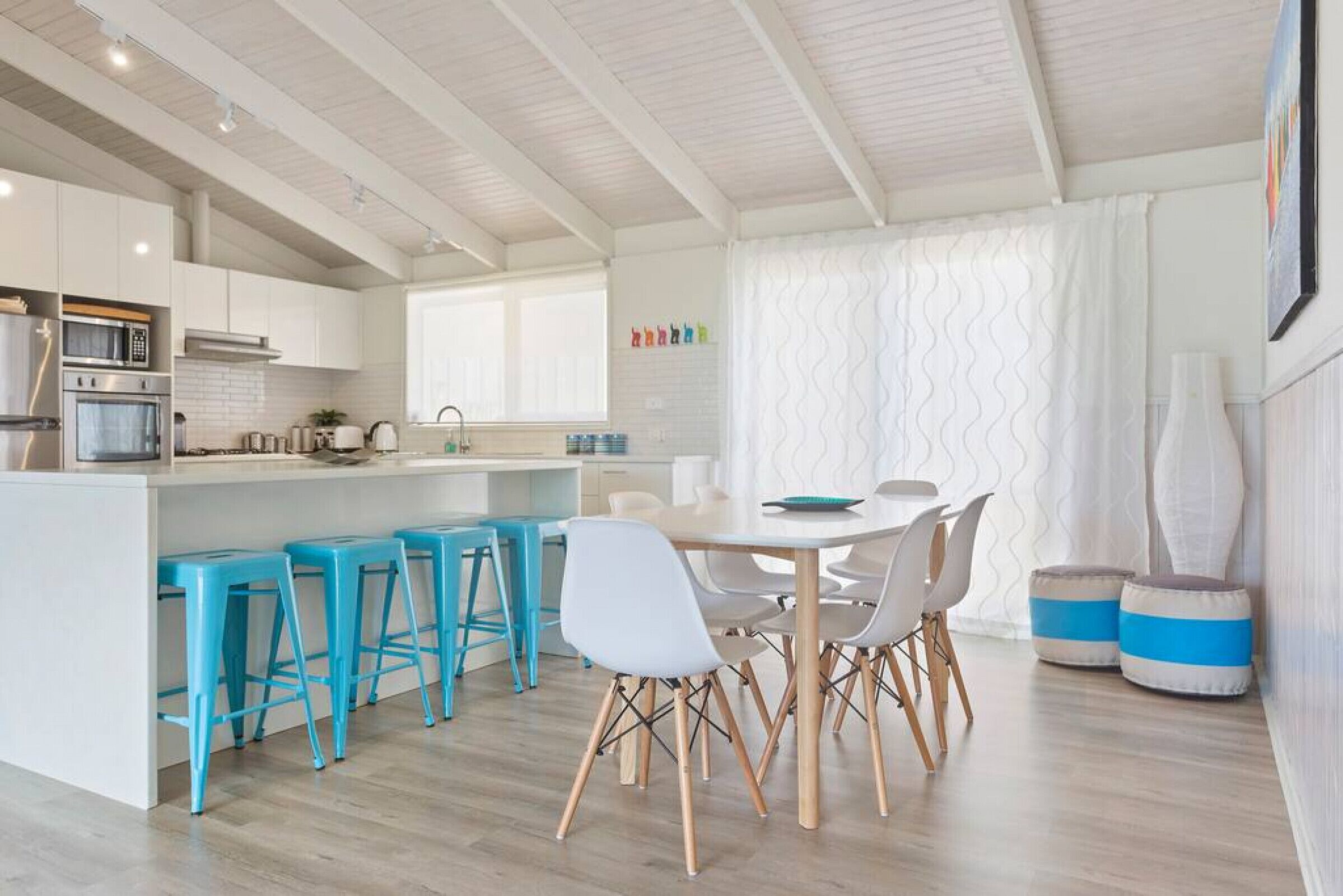 A modern kitchen with white cabinets, a built-in oven, and a microwave. There is a dining area with a white table and chairs, and a bar area with blue stools.
