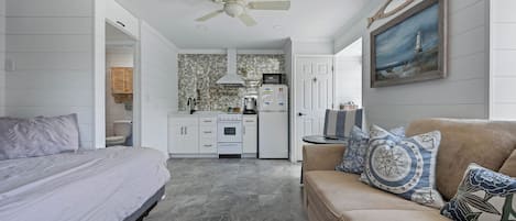 A bedroom with a bed, a sofa, a ceiling fan, and a kitchen in the background.