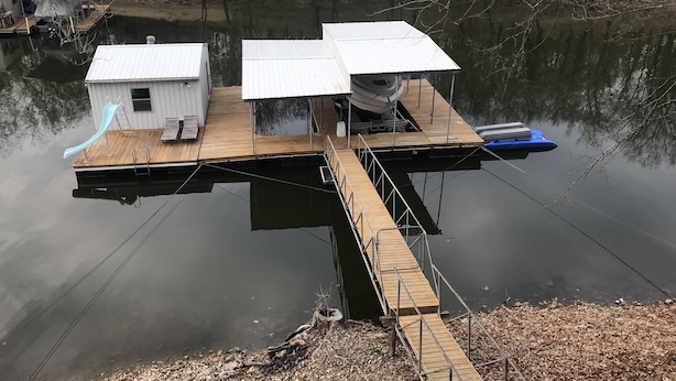 Une maison flottante avec un quai s’étendant sur un lac paisible, entourée d’arbres.