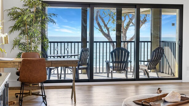 Una habitación con suelo de madera, una mesa de comedor con sillas, una planta en maceta y vista al mar a través de grandes puertas de cristal.