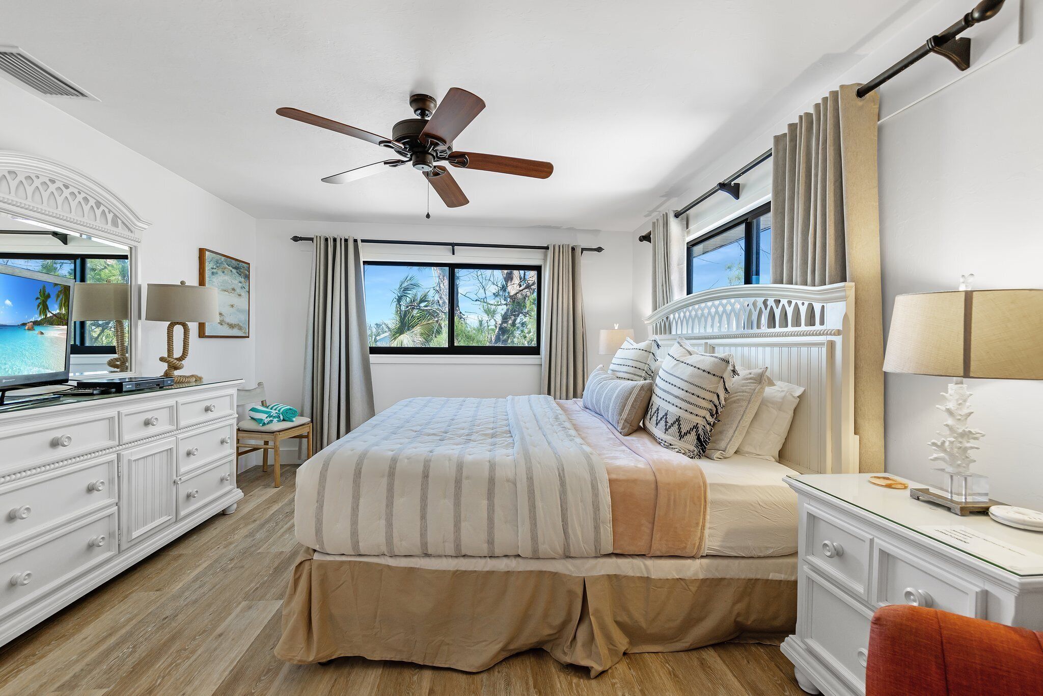 A bedroom with a large bed, a ceiling fan, a dresser, a nightstand, and a window with curtains.