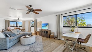 Una sala moderna con un sofá modular, una mesa de comedor con sillas, un ventilador de techo y un ventanal con vistas a árboles.