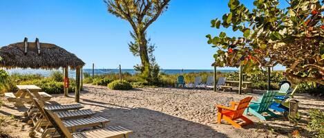 En strand med træloungestole og farverige strandstole, en stråtag parasol og havudsigt.