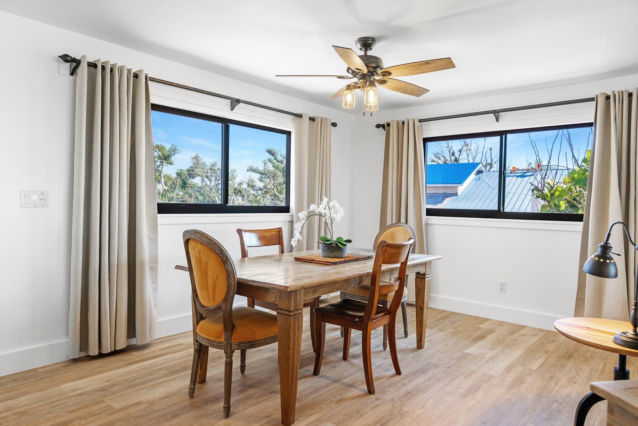 A dining room with a wooden table, four chairs, a ceiling fan, and large windows with curtains.