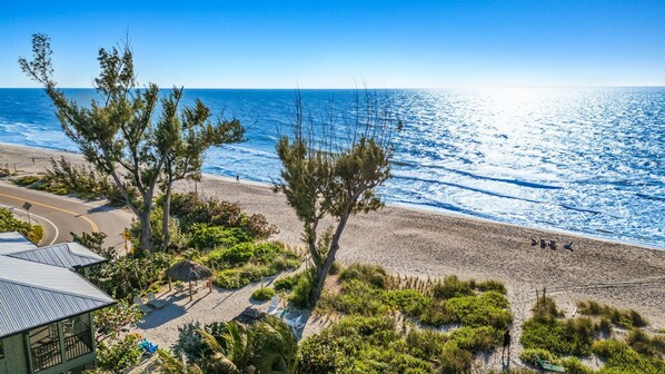 A beach with a clear view of the ocean, a few people walking, and a building with a thatched roof and outdoor seating.