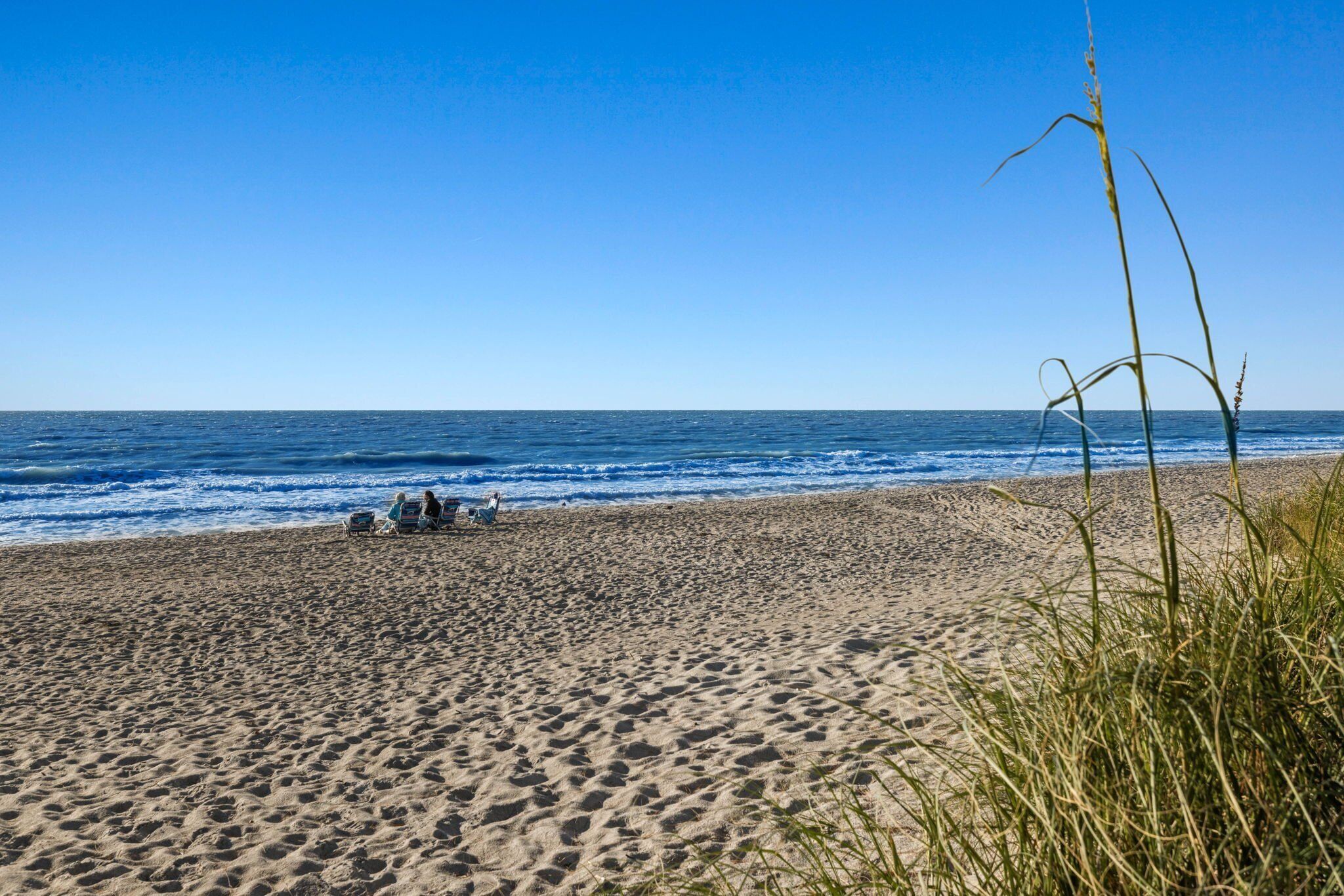 Une plage avec un ciel dégagé et une mer calme. On peut voir des gens assis sur le sable.