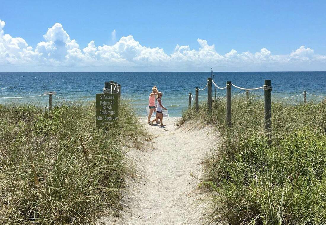 Un sentier sablonneux mène à une plage où un panneau indique qu’il faut rapporter tout le matériel de plage après utilisation.