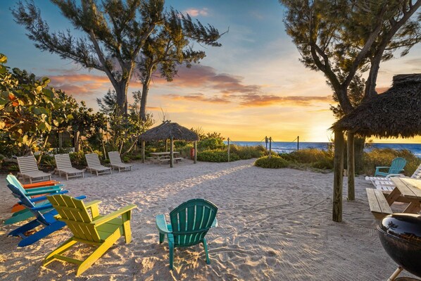 A beach setting with colorful Adirondack chairs, a thatched-roof structure, and a view of the ocean at sunset.